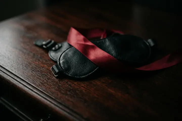 Bandeau en cuir noir et ruban de soie posés sur un bois sombre, sous une lumière rasante